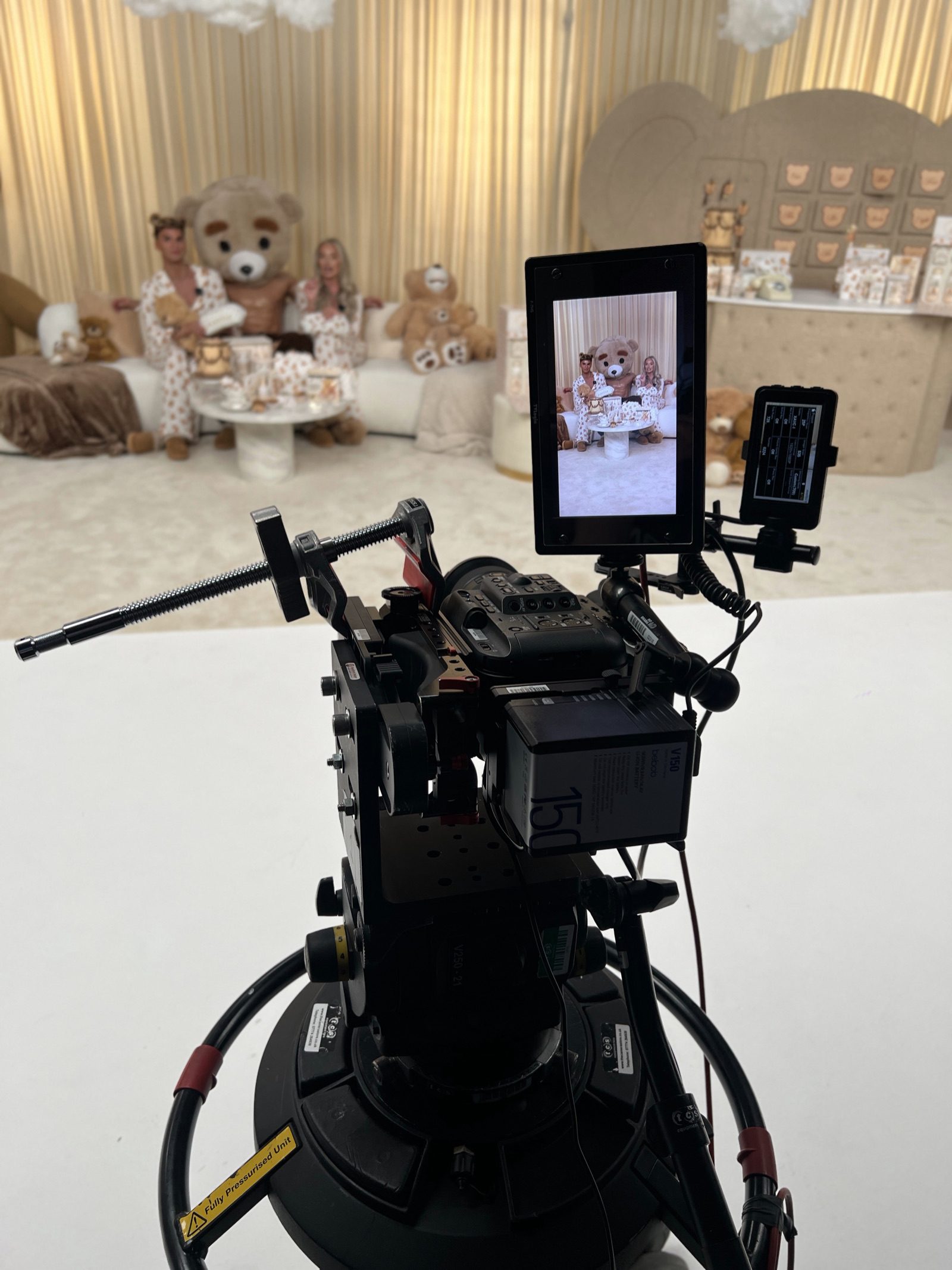 A broadcast camera and monitor pointed at two hosts on the P.Louise Teddy launch set, surrounded by giant teddy bears